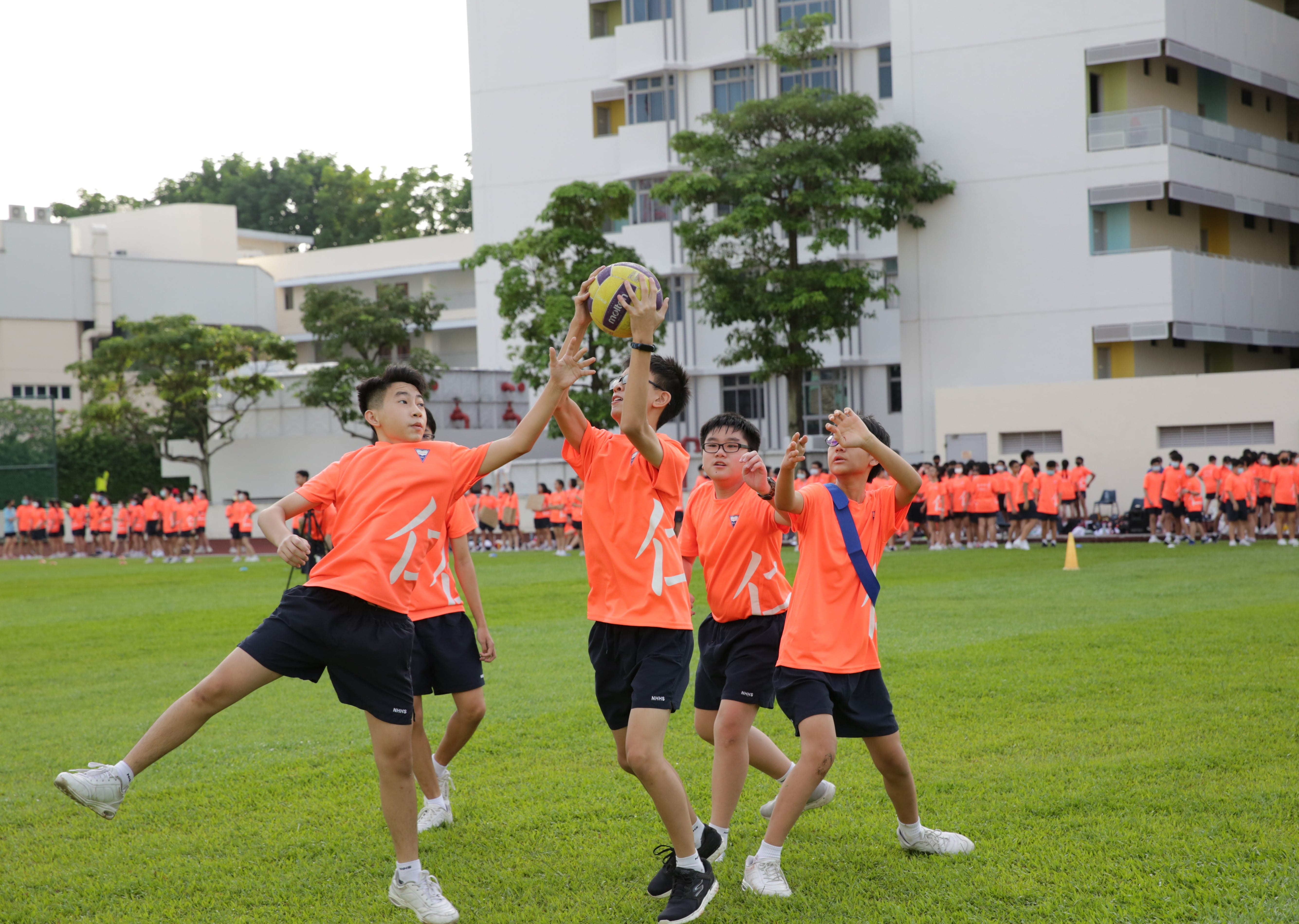 Four boys in orange jerseys compete for a ball on a grass field, with a crowd and school buildings behind them.